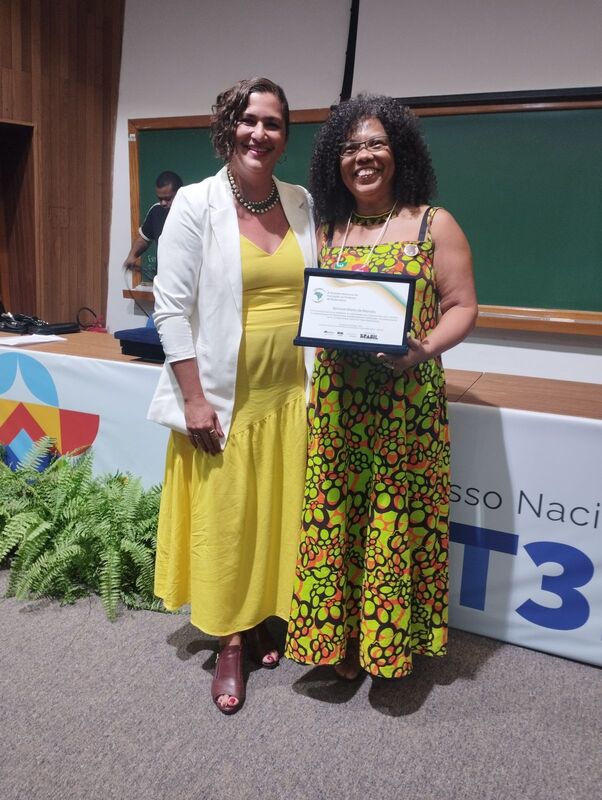Profa . Simone Moraes recebendo a homenagem das mãos de Tereza Farias, Coordenadora Geral do Ensino Fundamental do MEC.